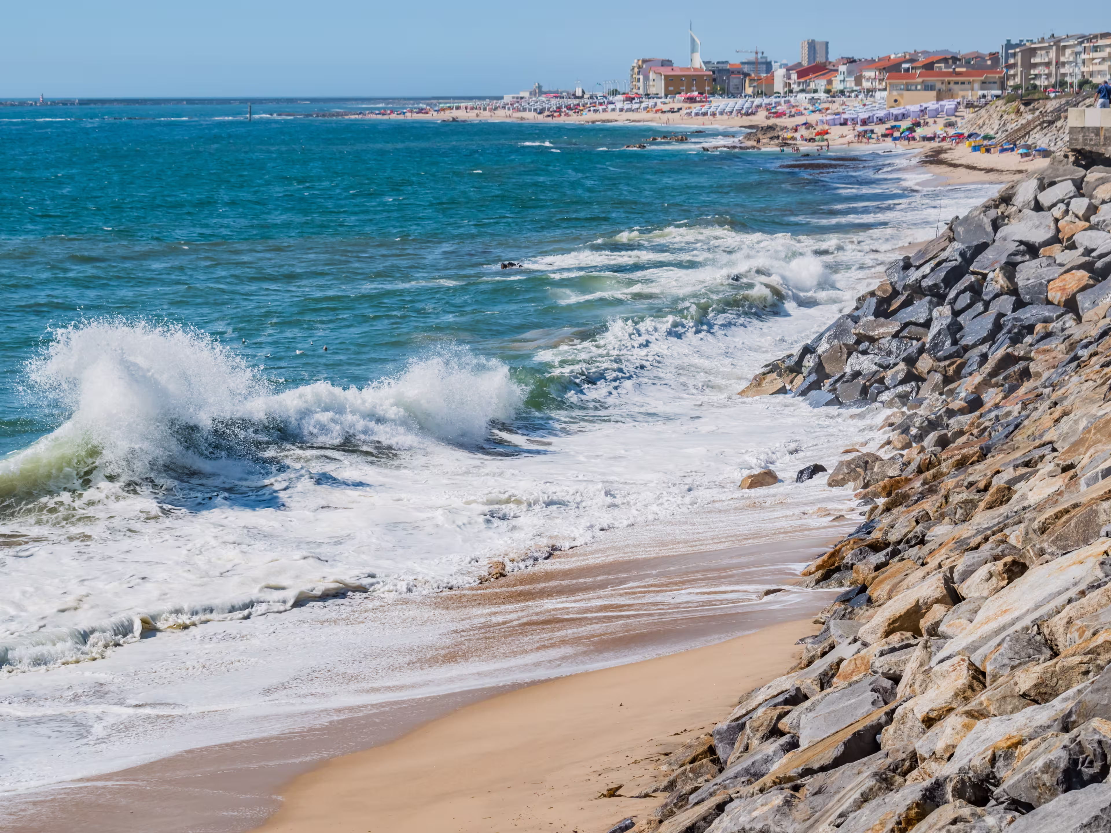 Selective focus of a wave on sand with a stone wall on Olinda beach with Póvoa de Varzim architecture breaking in blur in the background, Vila do Conde PORTUGAL