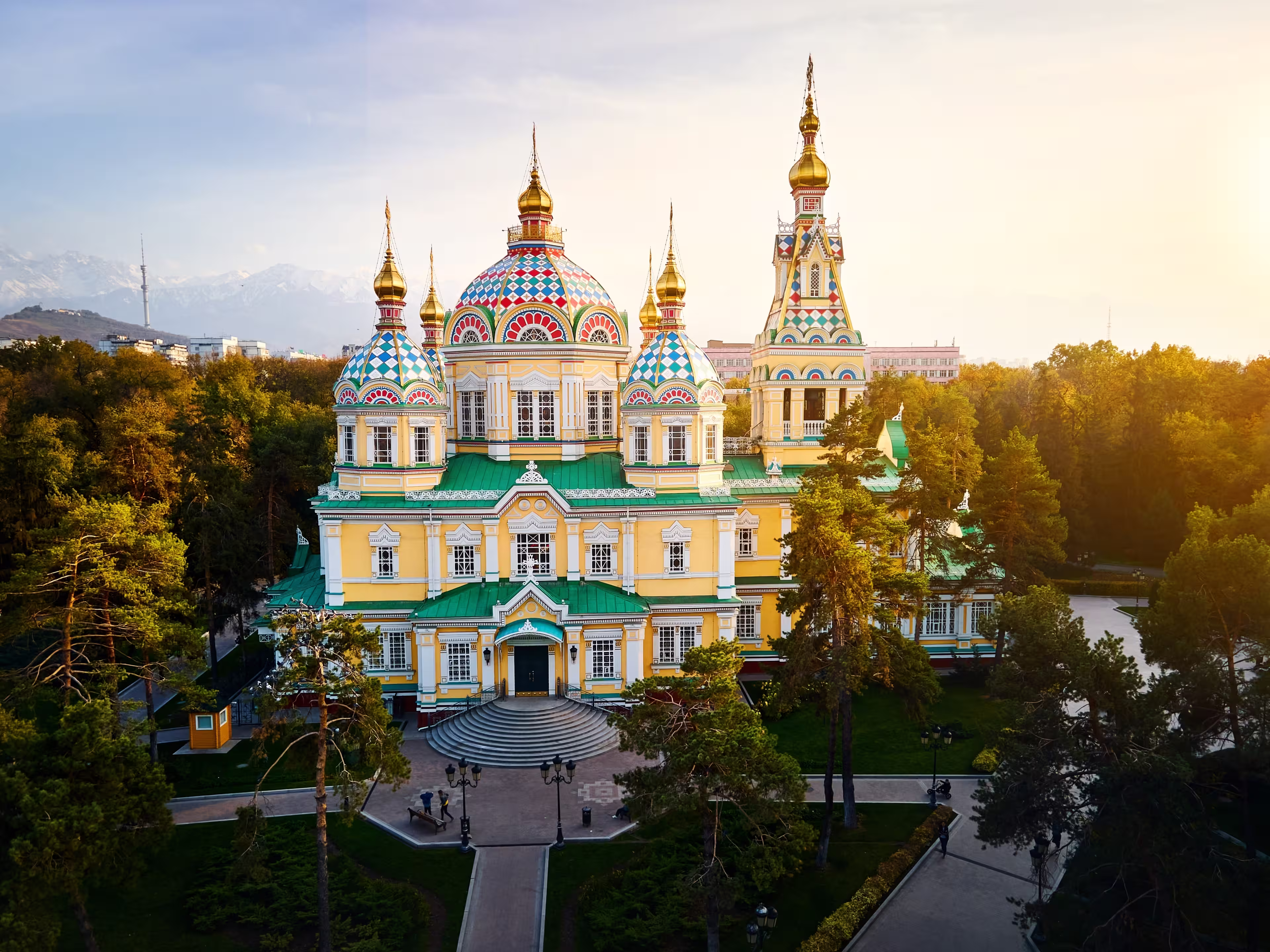 Aerial drone panorama of the Ascension Cathedral Russian Orthodox church and snow mountains at background in Panfilov Park against sunset sky in Almaty city, Kazakhstan