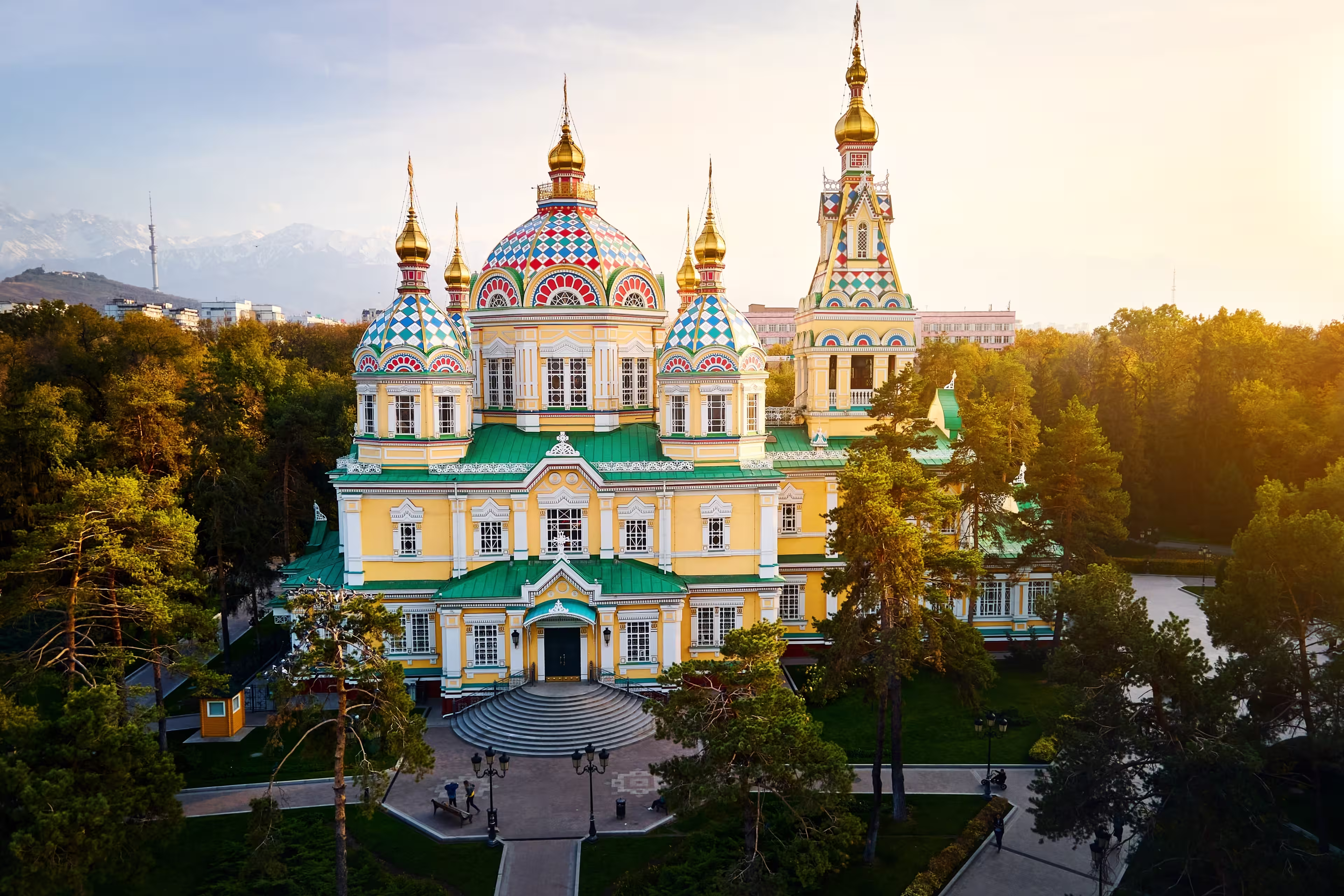 Aerial drone panorama of the Ascension Cathedral Russian Orthodox church and snow mountains at background in Panfilov Park against sunset sky in Almaty city, Kazakhstan
