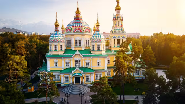 Aerial drone panorama of the Ascension Cathedral Russian Orthodox church and snow mountains at background in Panfilov Park against sunset sky in Almaty city, Kazakhstan