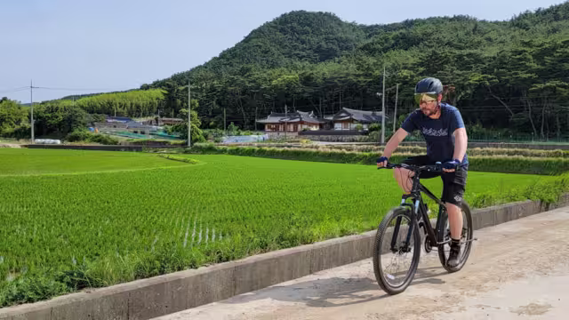 Cyclist in helmet and gear cycling in rural South Korea past traditional houses and fields