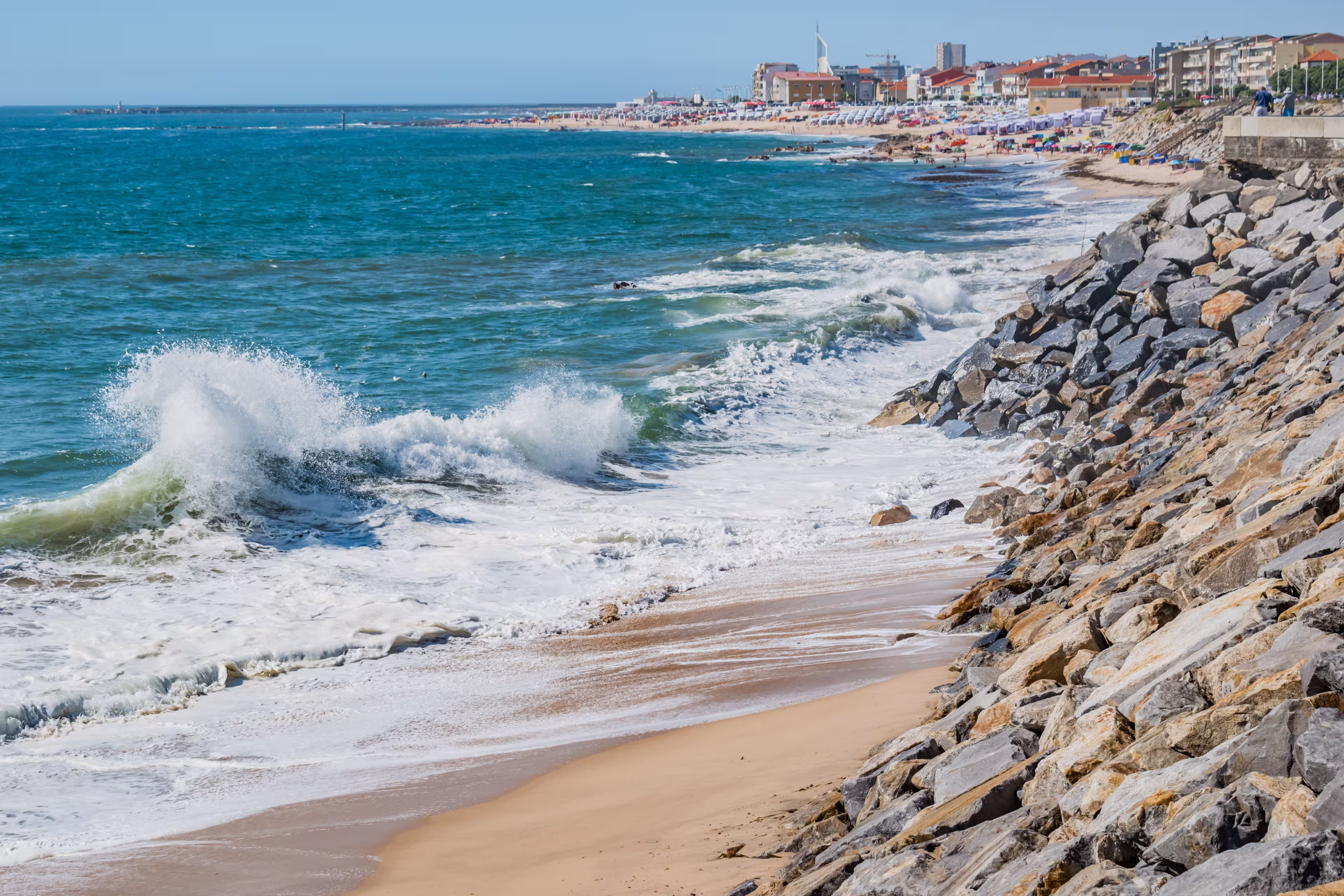 Selective focus of a wave on sand with a stone wall on Olinda beach with Póvoa de Varzim architecture breaking in blur in the background, Vila do Conde PORTUGAL