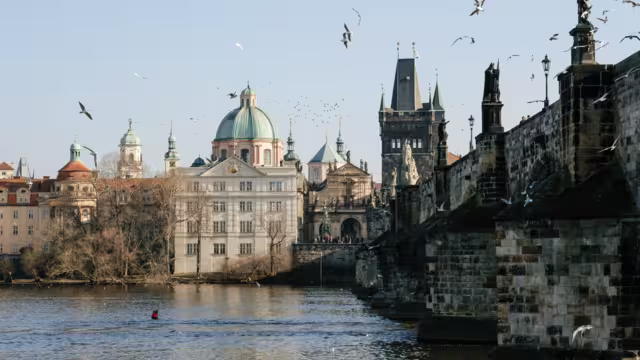 Historic Charles Bridge with Gothic towers spans Vltava River under vibrant blue February sky. Famous Prague landmark with baroque statues glowing in winter sunlight.