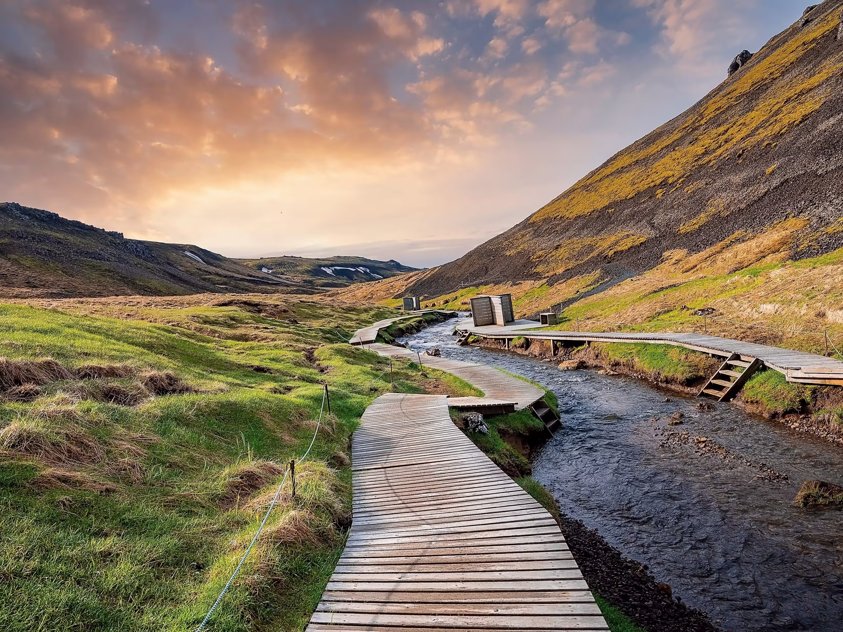 Beautiful view of wooden boardwalk by hot river stream. Natural geothermal bath in Reykjadalur Valley against cloudy sky. Grassy landscape by mountains in Hveragerdi during sunset.