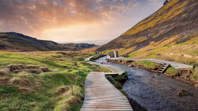 Beautiful view of wooden boardwalk by hot river stream. Natural geothermal bath in Reykjadalur Valley against cloudy sky. Grassy landscape by mountains in Hveragerdi during sunset.
