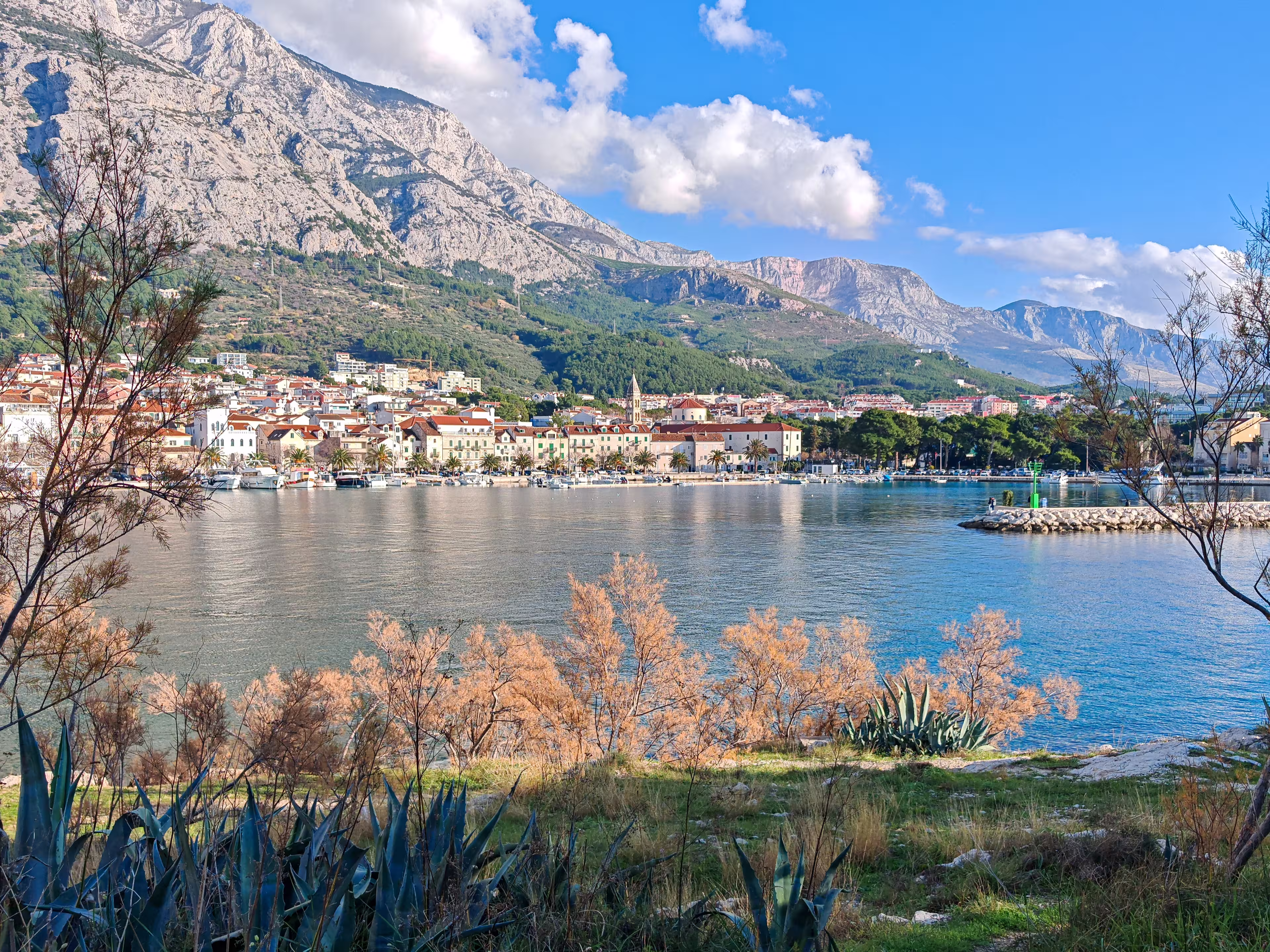 Autumn view of Makarska set between the calm Adriatic Sea and the rugged Biokovo mountains, where coastal architecture meets dramatic limestone slopes under soft seasonal light.