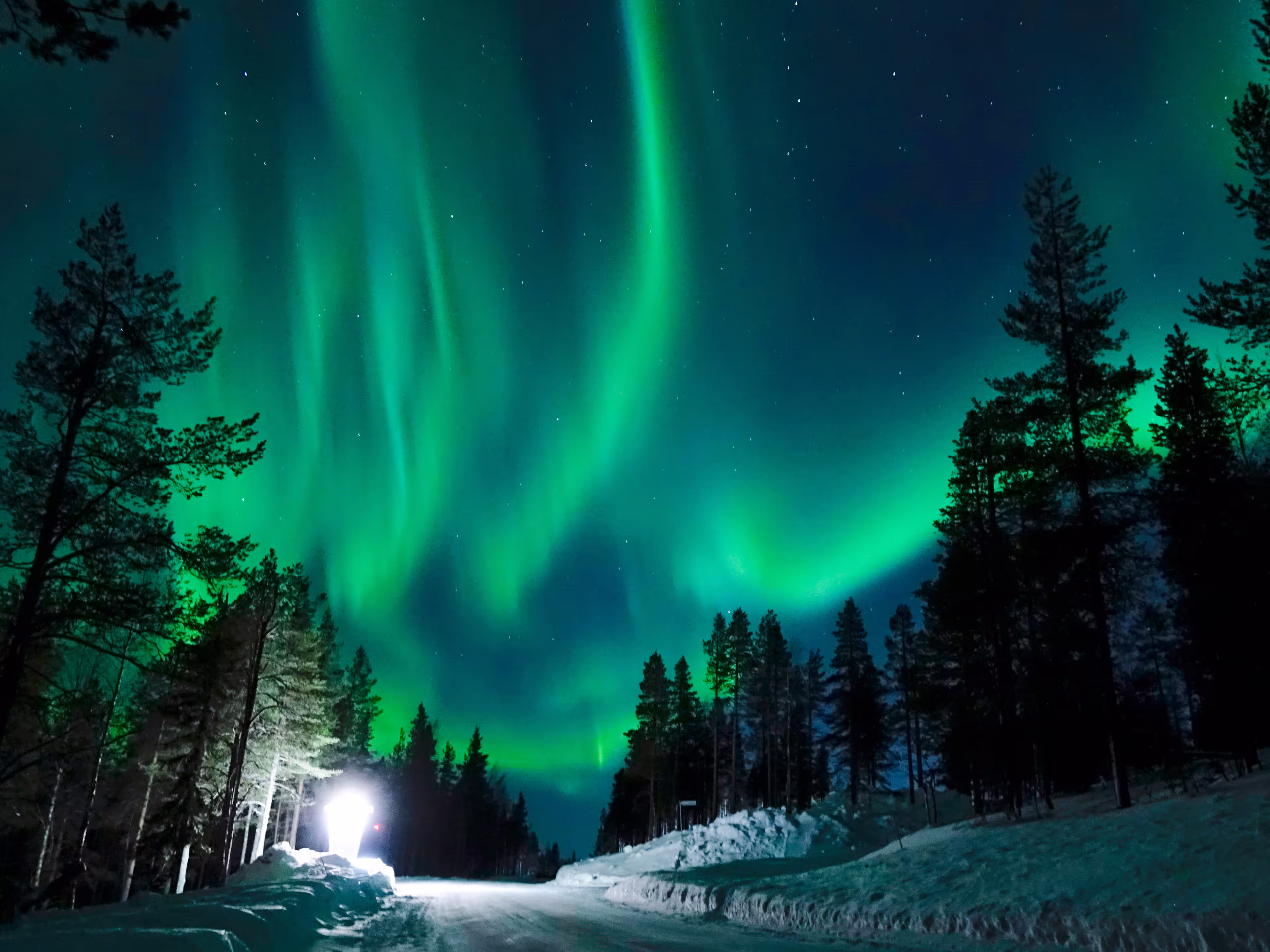 Bright lamp shining on the empty snowy road just as the northern lights appear above the idyllic Scandinavian countryside in the winter. Breathtaking aurora borealis above the calm wintry landscape.