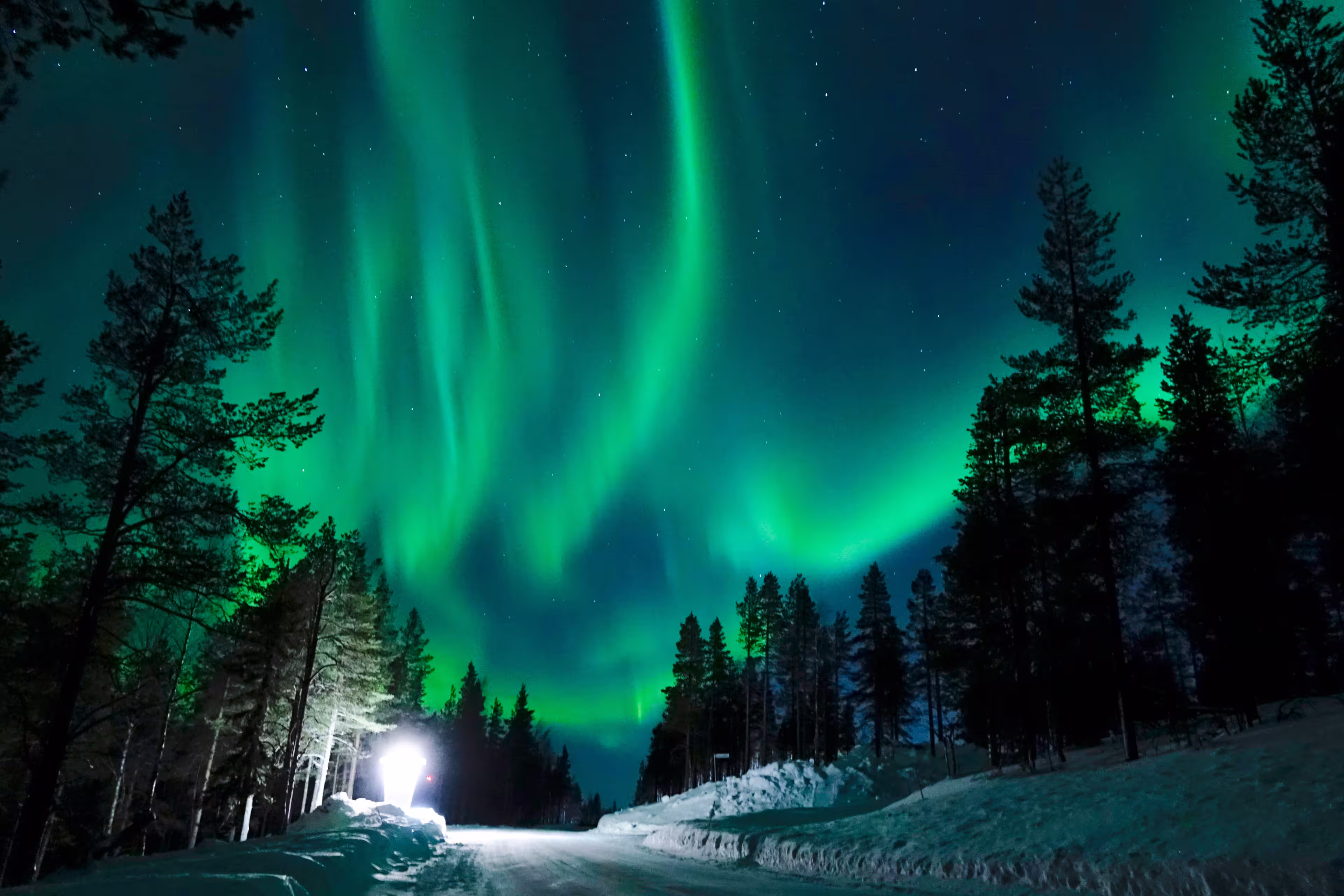 Bright lamp shining on the empty snowy road just as the northern lights appear above the idyllic Scandinavian countryside in the winter. Breathtaking aurora borealis above the calm wintry landscape.