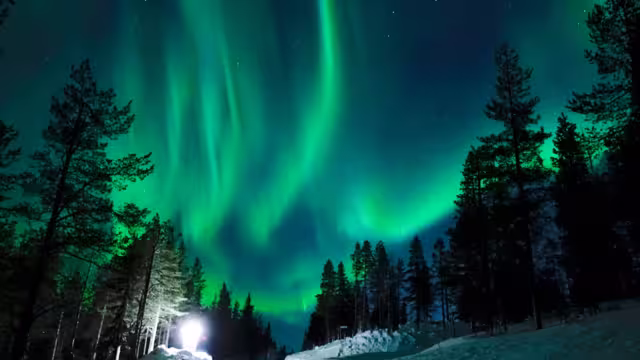 Bright lamp shining on the empty snowy road just as the northern lights appear above the idyllic Scandinavian countryside in the winter. Breathtaking aurora borealis above the calm wintry landscape.