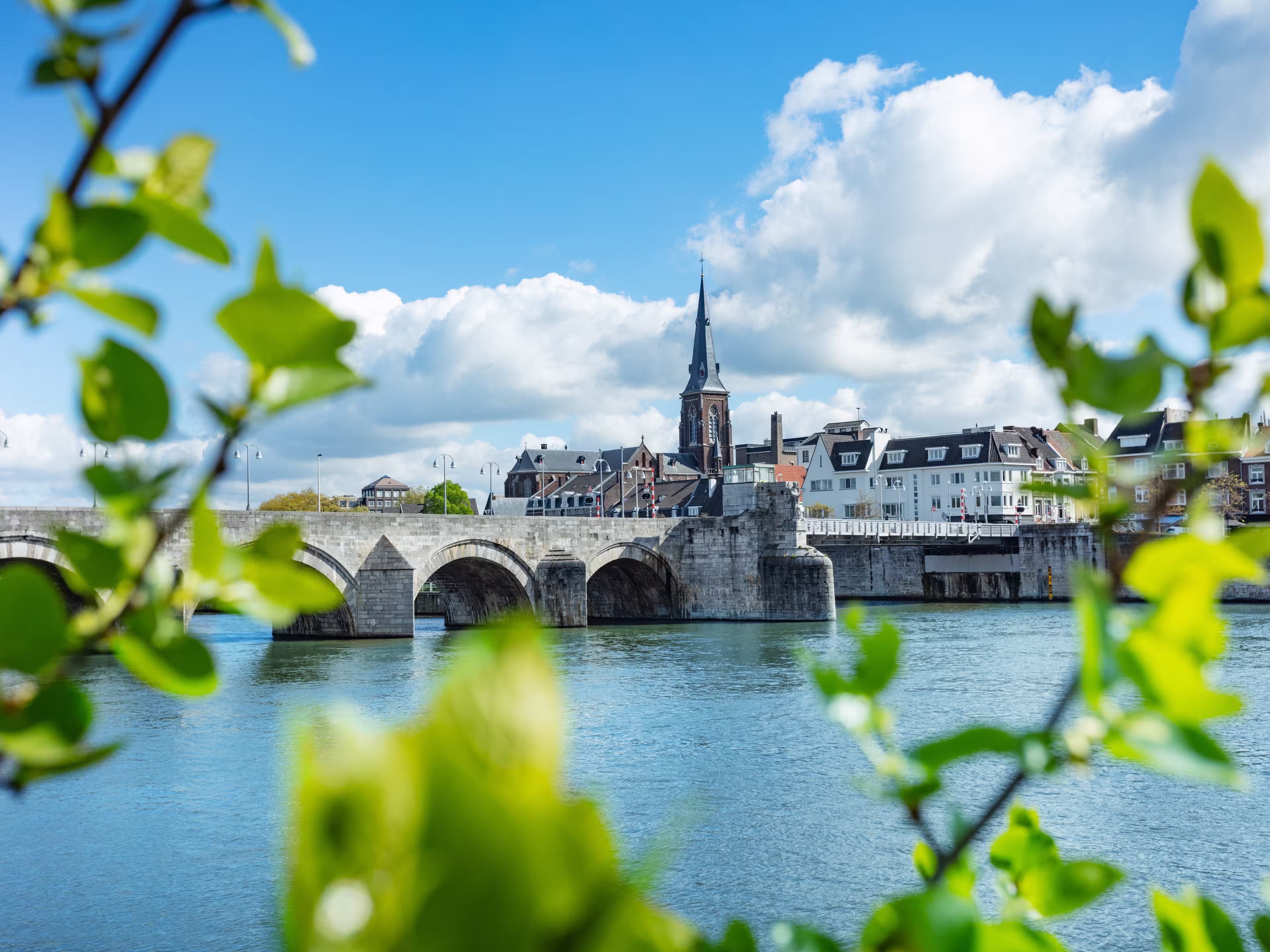 Historic stone Sint Servaas bridge spanning across a serene river, framed by fresh green foliage under a bright blue sky, Maastricht, Holland