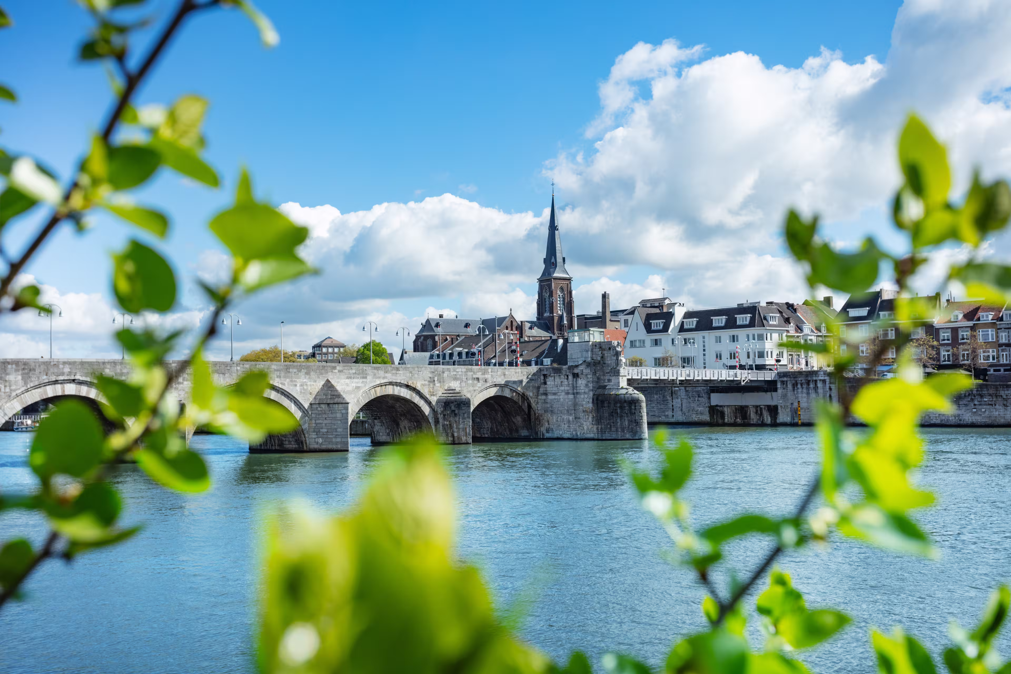 Historic stone Sint Servaas bridge spanning across a serene river, framed by fresh green foliage under a bright blue sky, Maastricht, Holland