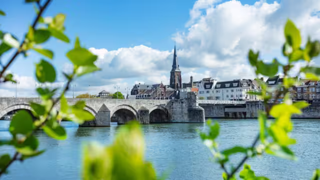 Historic stone Sint Servaas bridge spanning across a serene river, framed by fresh green foliage under a bright blue sky, Maastricht, Holland
