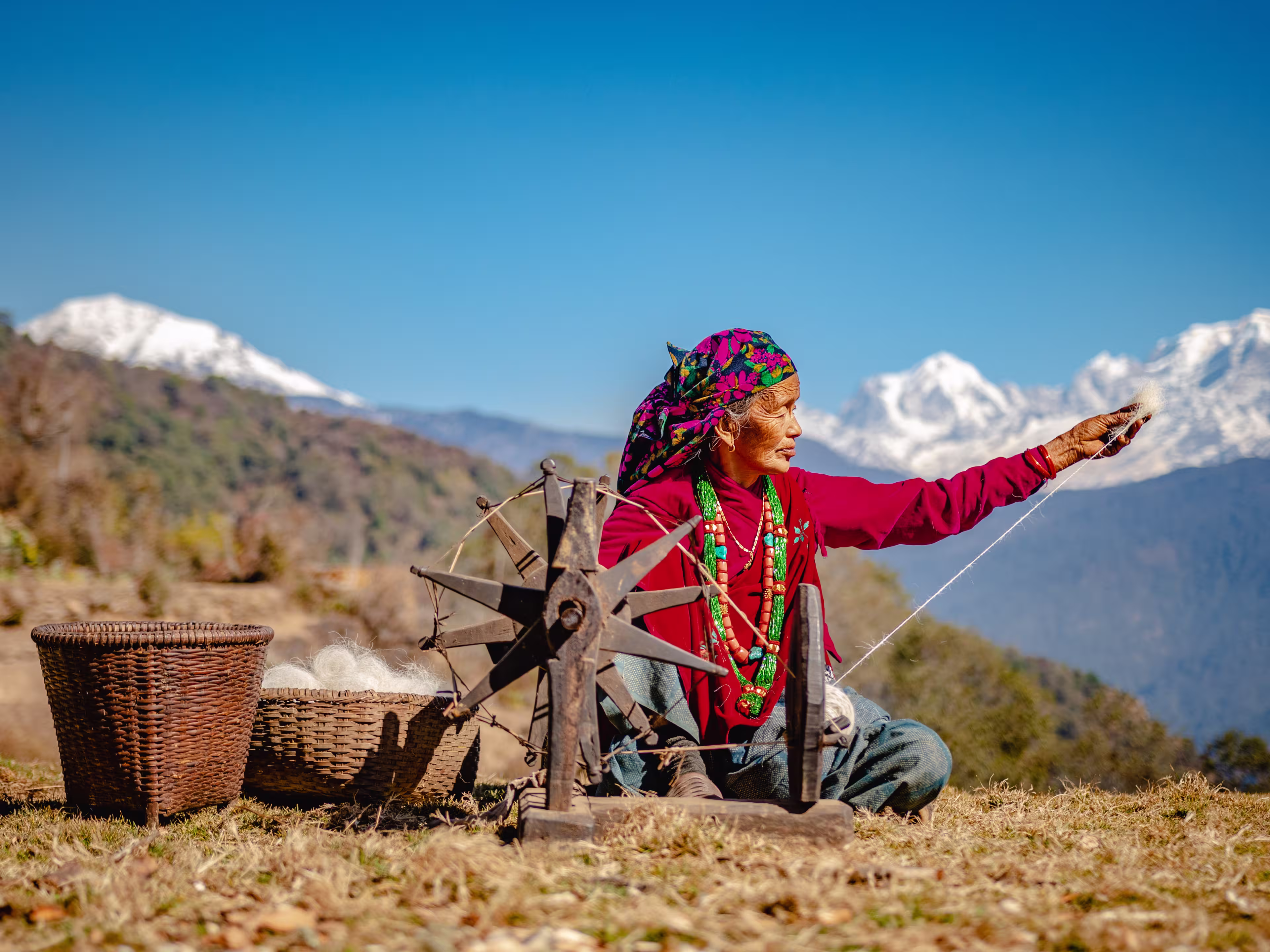 GHAN POKHARA, GANDAKI/NEPAL - 02/03/2019: A portrait of an elderly Nepali Gurung woman in her traditional dress weaving cotton from wool on a sunny day with Annapurna Mountain Range in the background.