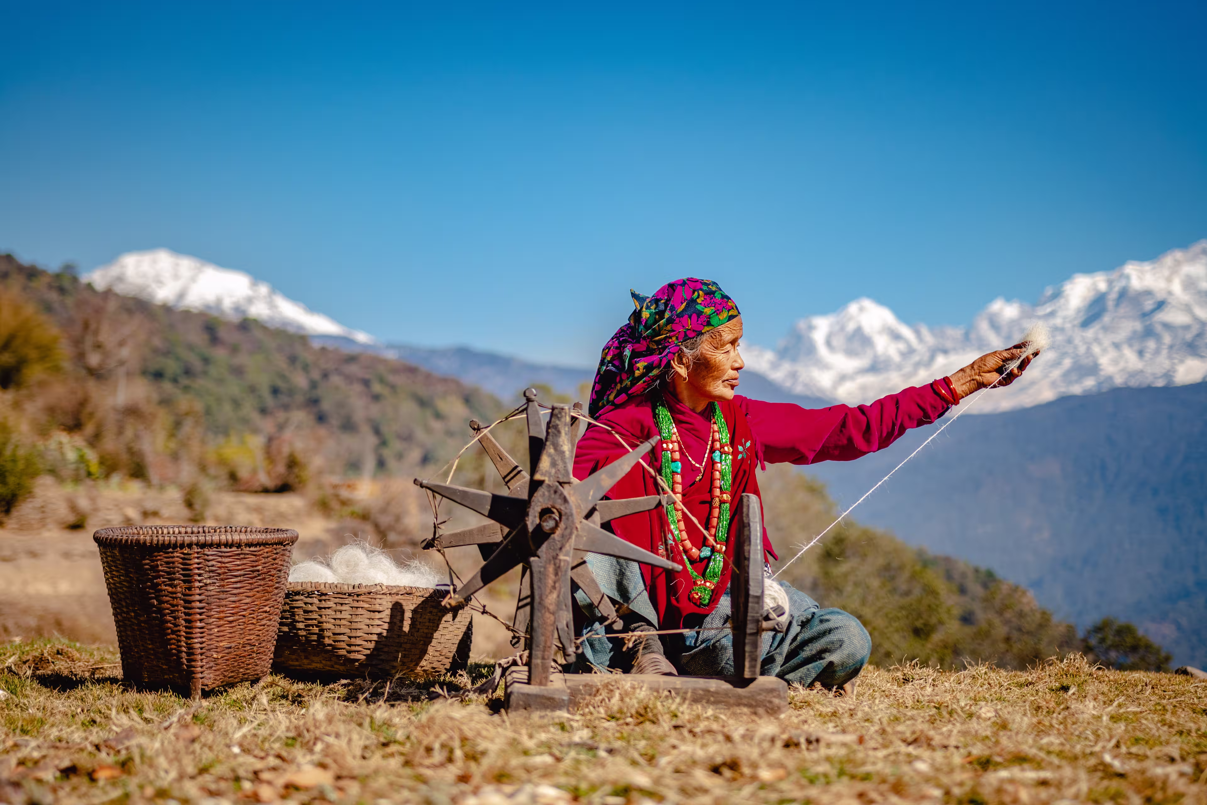 GHAN POKHARA, GANDAKI/NEPAL - 02/03/2019: A portrait of an elderly Nepali Gurung woman in her traditional dress weaving cotton from wool on a sunny day with Annapurna Mountain Range in the background.