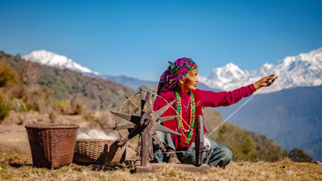 GHAN POKHARA, GANDAKI/NEPAL - 02/03/2019: A portrait of an elderly Nepali Gurung woman in her traditional dress weaving cotton from wool on a sunny day with Annapurna Mountain Range in the background.