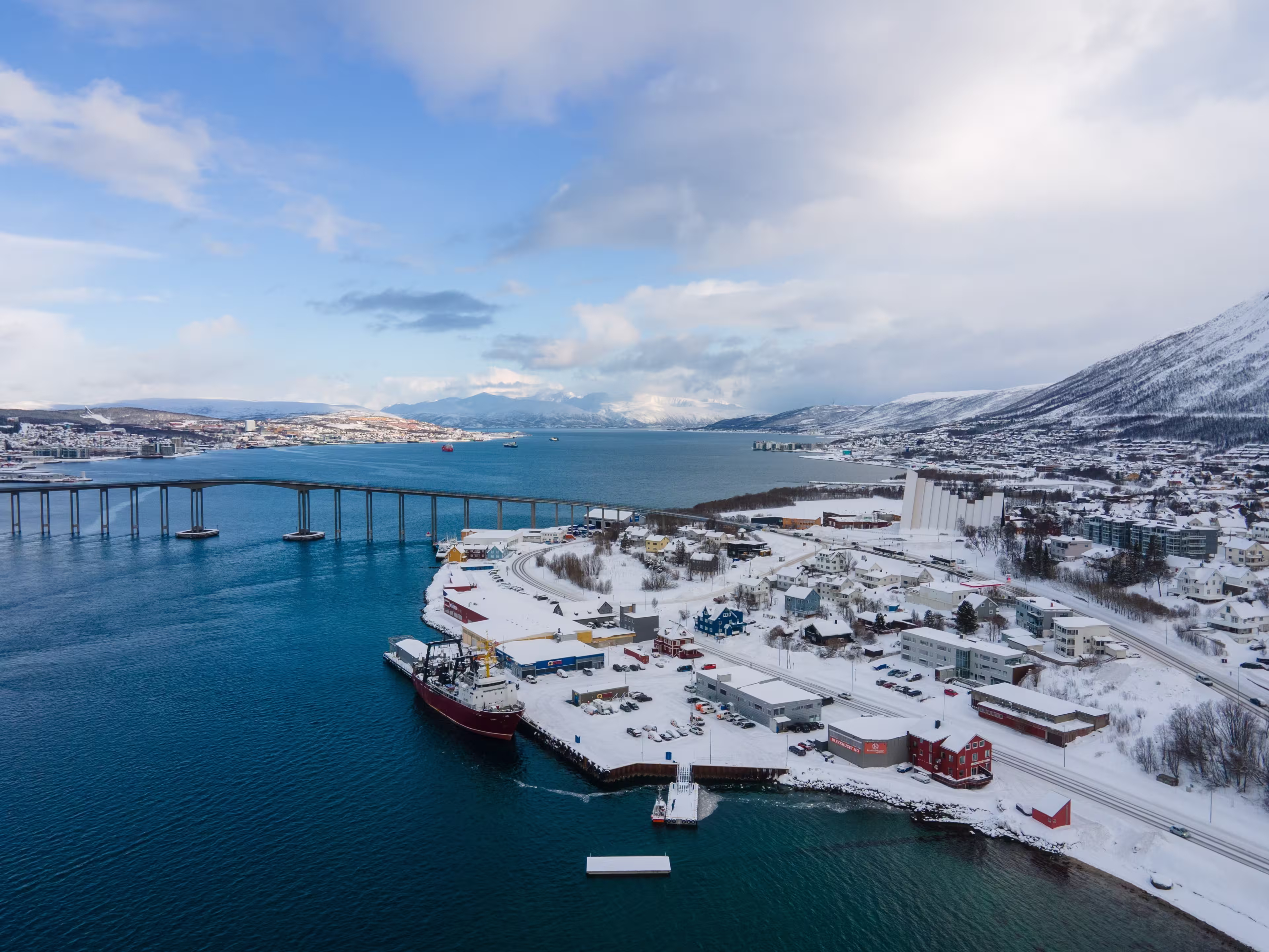 Drone shot of Tromsø in winter, showcasing the iconic Arctic Cathedral, Tromsø Bridge, snowy landscape, and surrounding waters under a clear blue Arctic sky.