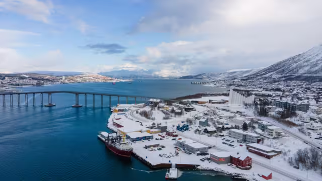 Drone shot of Tromsø in winter, showcasing the iconic Arctic Cathedral, Tromsø Bridge, snowy landscape, and surrounding waters under a clear blue Arctic sky.