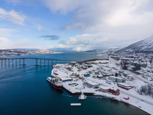 Drone shot of Tromsø in winter, showcasing the iconic Arctic Cathedral, Tromsø Bridge, snowy landscape, and surrounding waters under a clear blue Arctic sky.