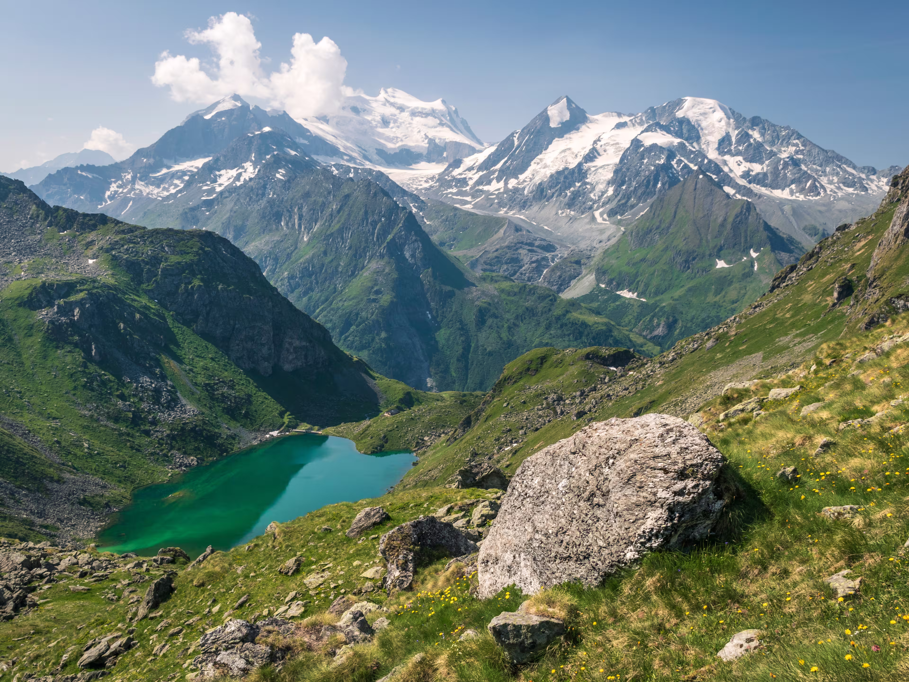 Turquoise Lac de Louvie in Swiss Alps, seen from above, with green meadows in the foreground and majestic, snowy Grand Combin massif in the background. The photo was taken in sunny, summer conditions.