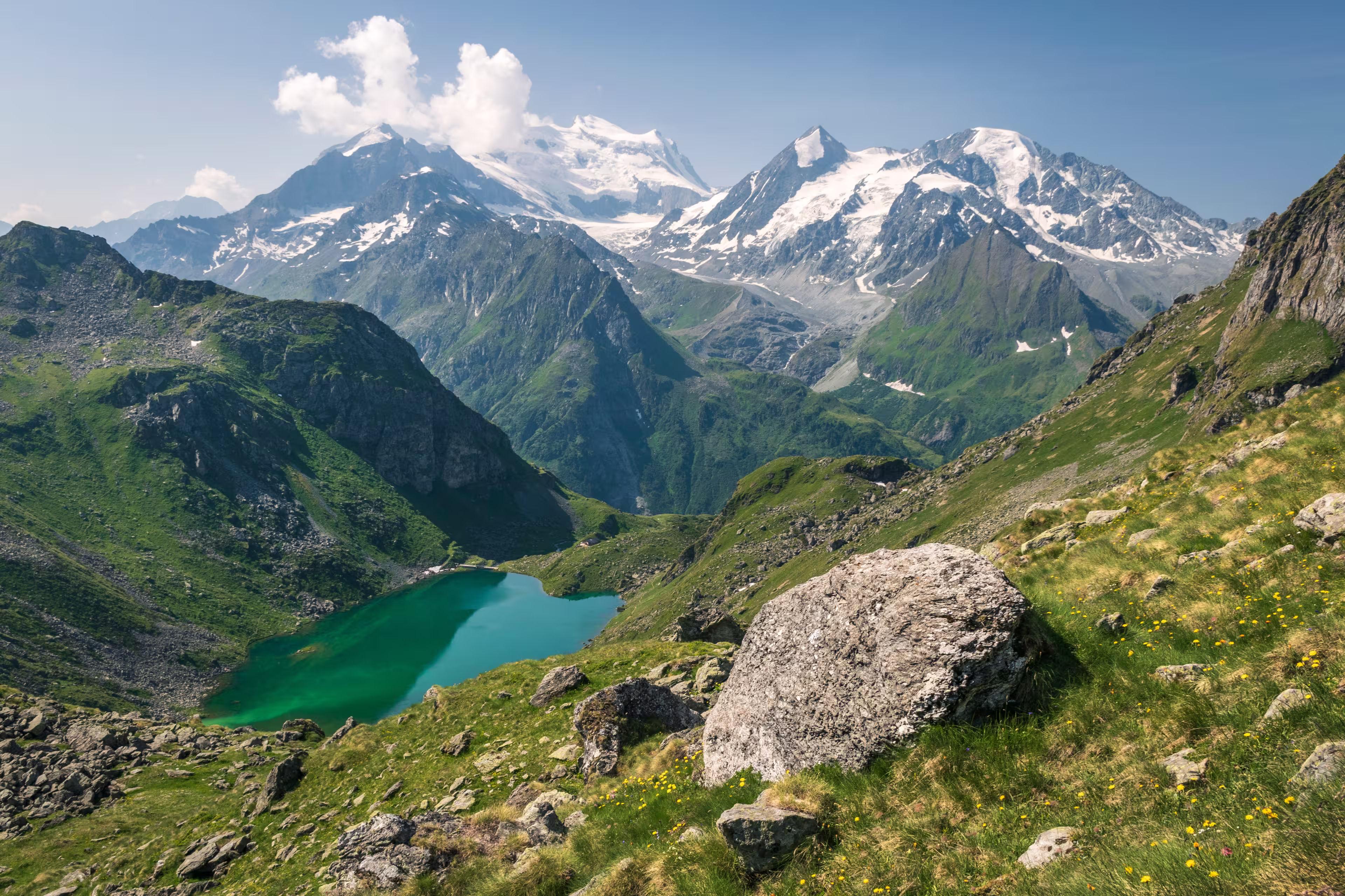 Turquoise Lac de Louvie in Swiss Alps, seen from above, with green meadows in the foreground and majestic, snowy Grand Combin massif in the background. The photo was taken in sunny, summer conditions.