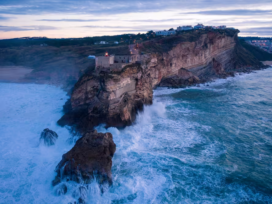 Sunset drone shot from the sea with waves breaking on the rocks in the foreground and Nazaré lighthouse and landscape with lights of the village in the background