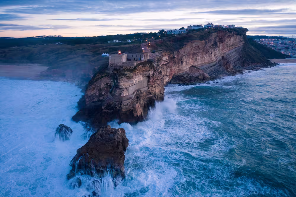Sunset drone shot from the sea with waves breaking on the rocks in the foreground and Nazaré lighthouse and landscape with lights of the village in the background