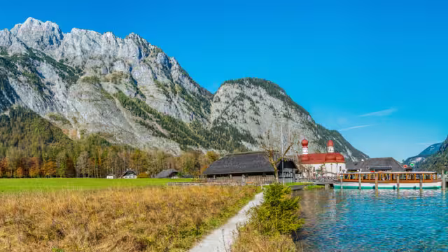 Panorama image of the Koenigssee with the chapel of st bartholomew and a tourist boat. In the background the illuminated mountain formation of Watzmann in bavaria, Germany