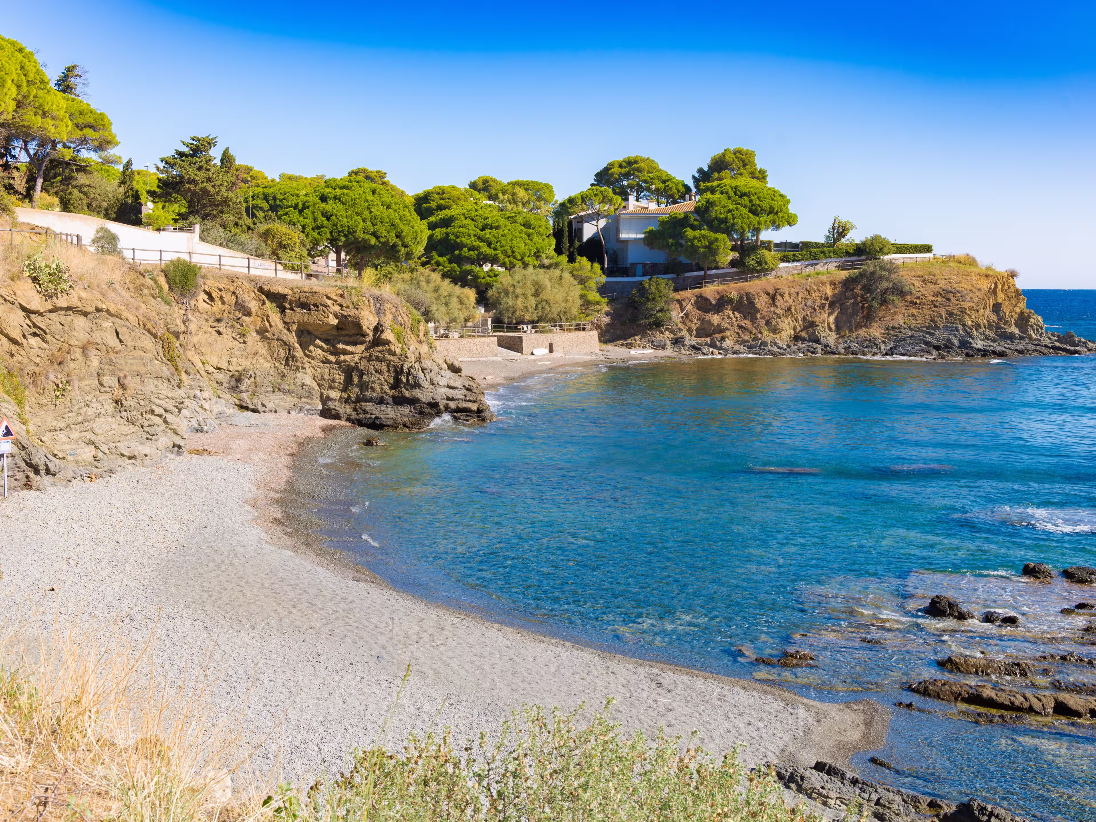 Quiet pebble beach at La Farella along the coastal path from Llançà to Port de la Selva, with turquoise waters, rocky cliffs and lush Mediterranean trees.