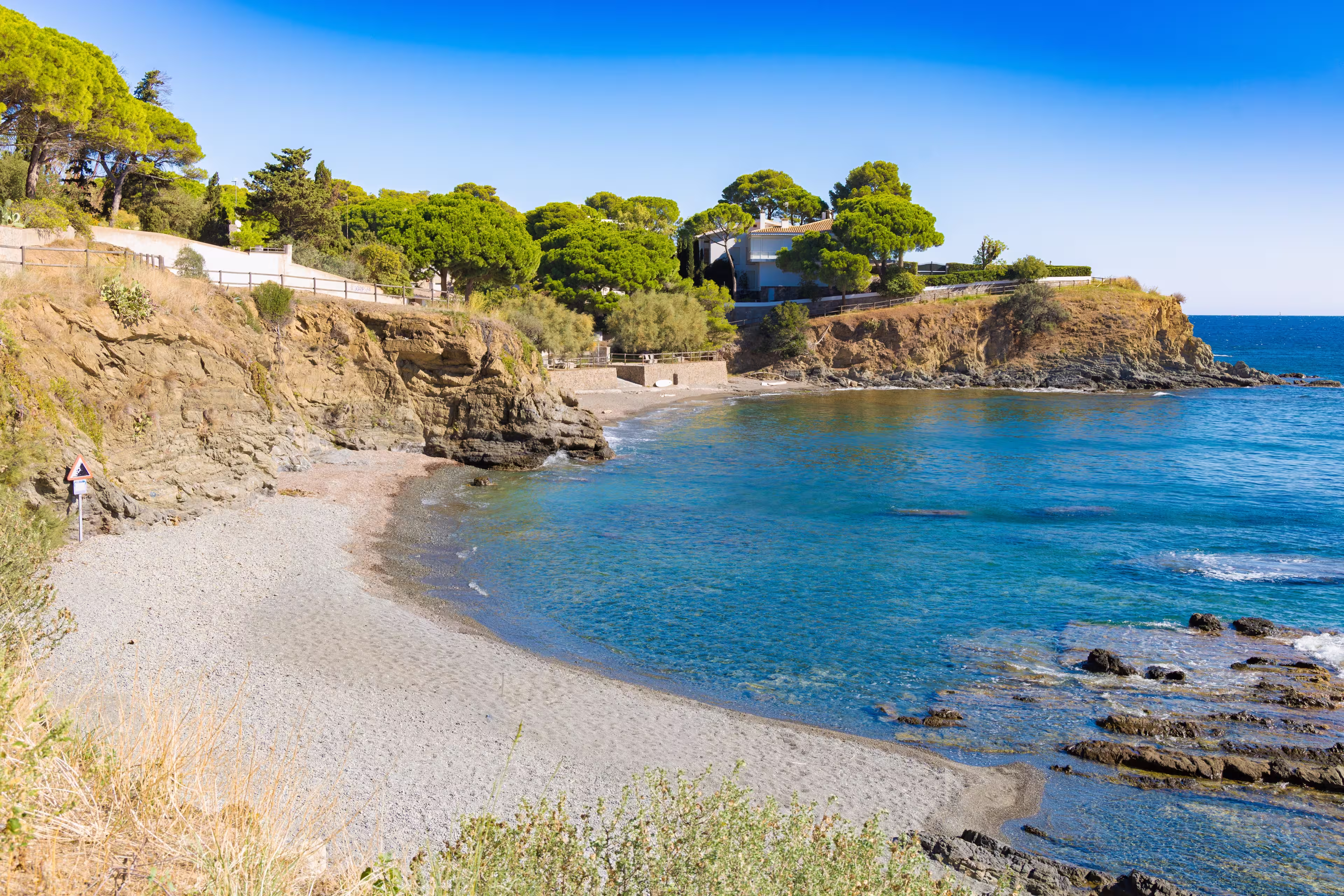 Quiet pebble beach at La Farella along the coastal path from Llançà to Port de la Selva, with turquoise waters, rocky cliffs and lush Mediterranean trees.