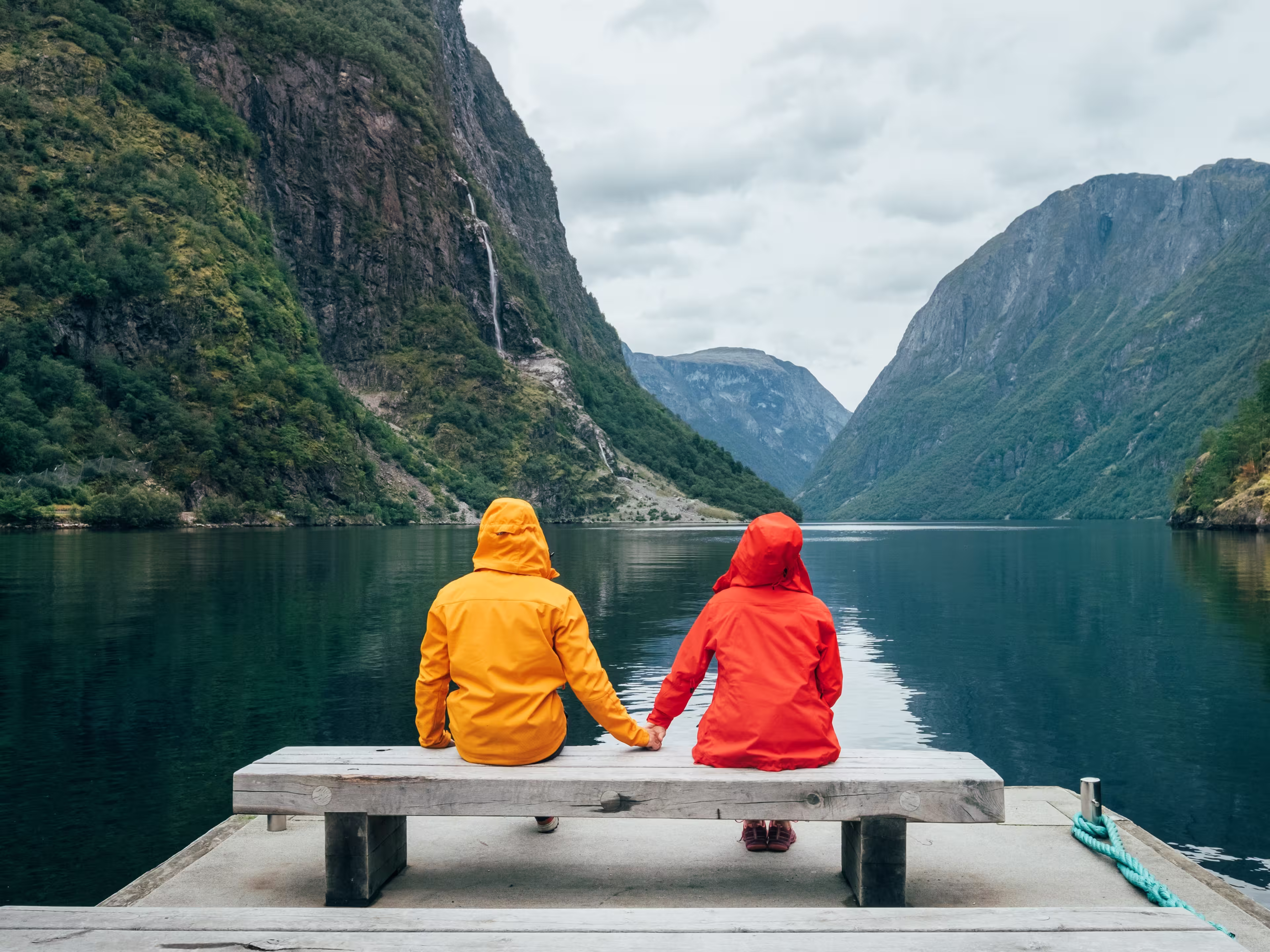Happy couple dressed in bright windproof jackets sitting and hugging on bench on the Norwegian Nærøyfjord bank in Gudvangen, Norway. Traveling, beauty in Nature and relations concept.