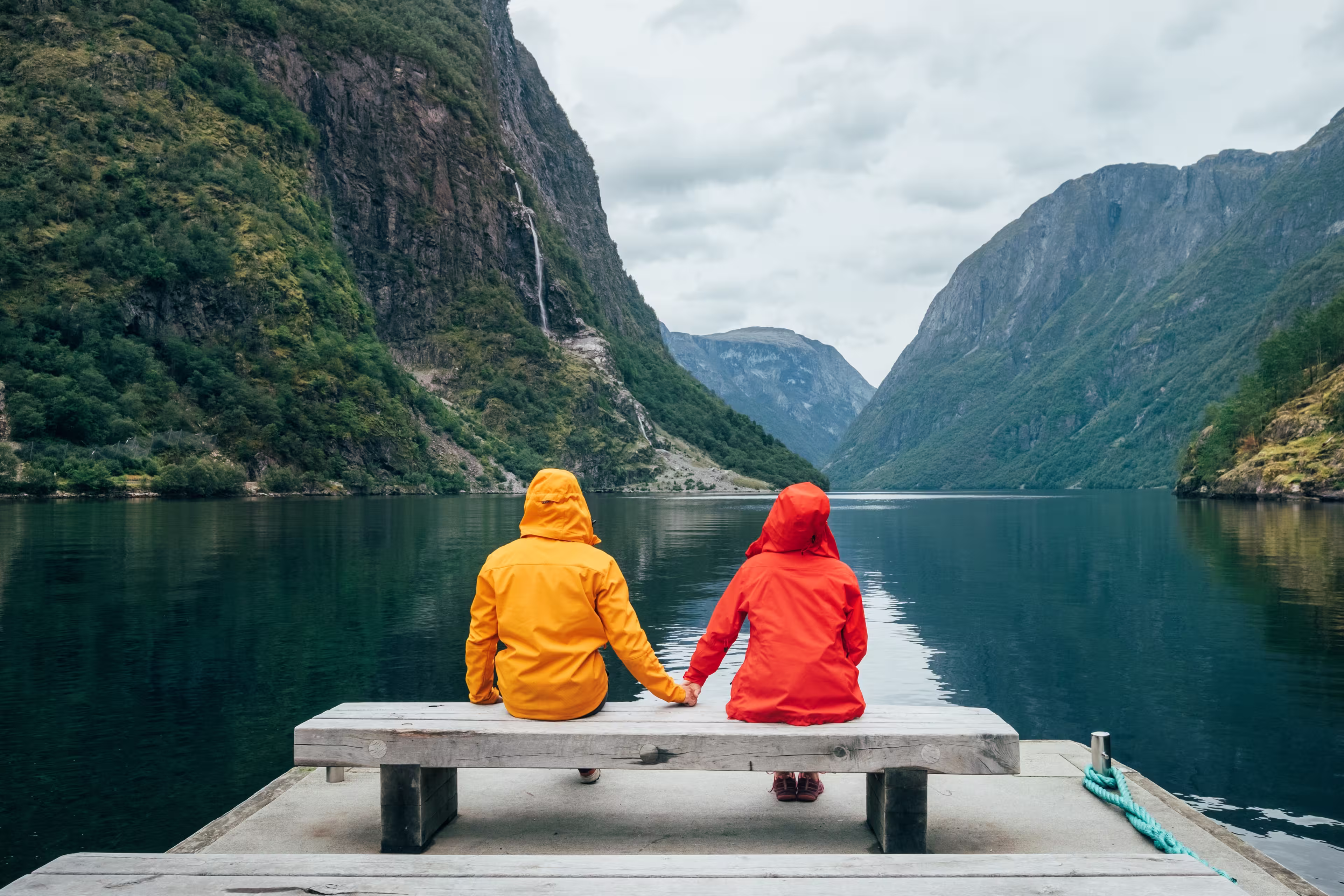 Happy couple dressed in bright windproof jackets sitting and hugging on bench on the Norwegian Nærøyfjord bank in Gudvangen, Norway. Traveling, beauty in Nature and relations concept.