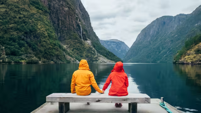 Happy couple dressed in bright windproof jackets sitting and hugging on bench on the Norwegian Nærøyfjord bank in Gudvangen, Norway. Traveling, beauty in Nature and relations concept.