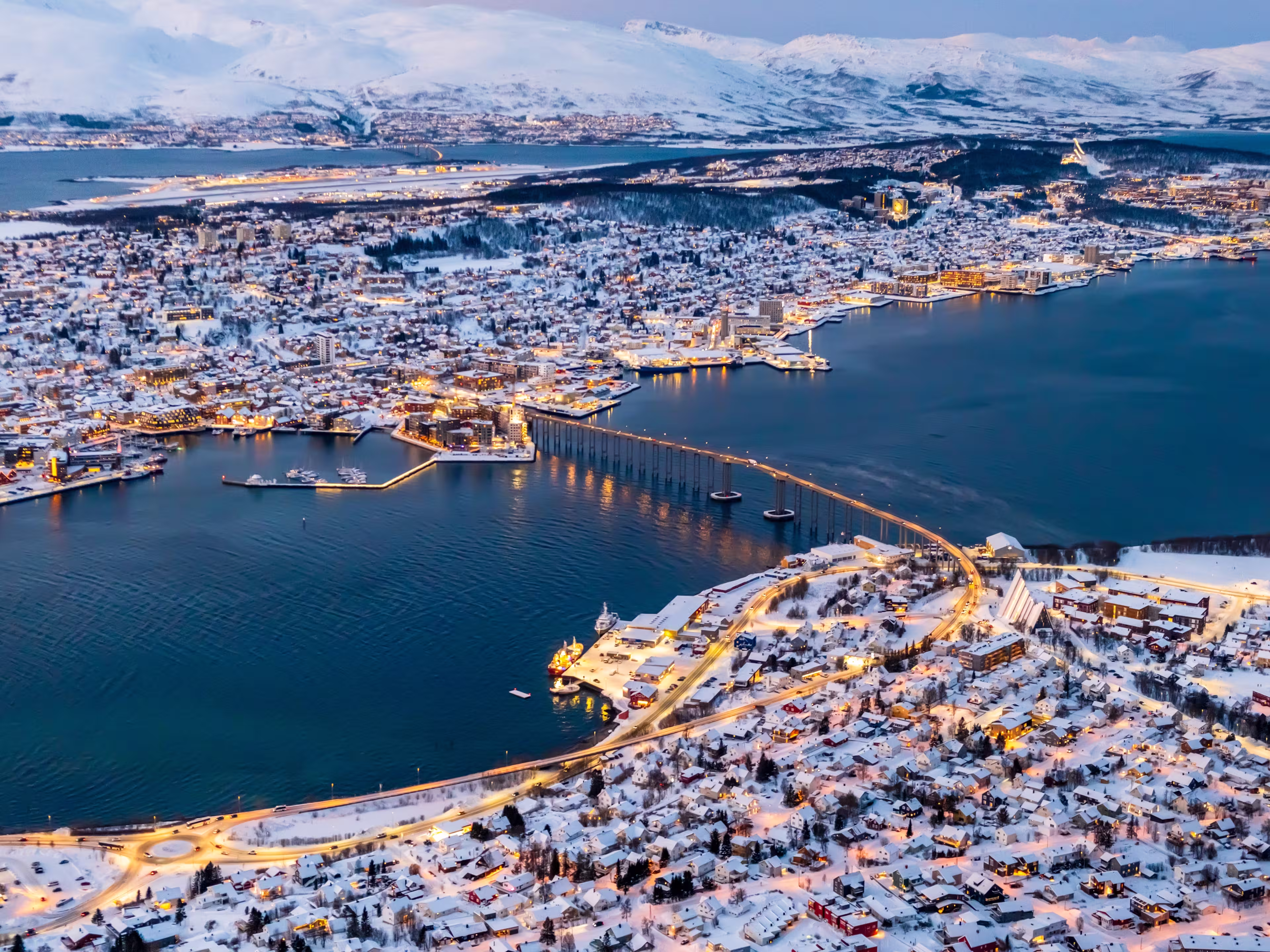 Aerial view of Tromso, Norway with city lights in winter from Storsteinen mountain. Tromsoya island with snowy roofs of Tromso town, harbor, Sandnessundet bridge over Fjord and mountains in background