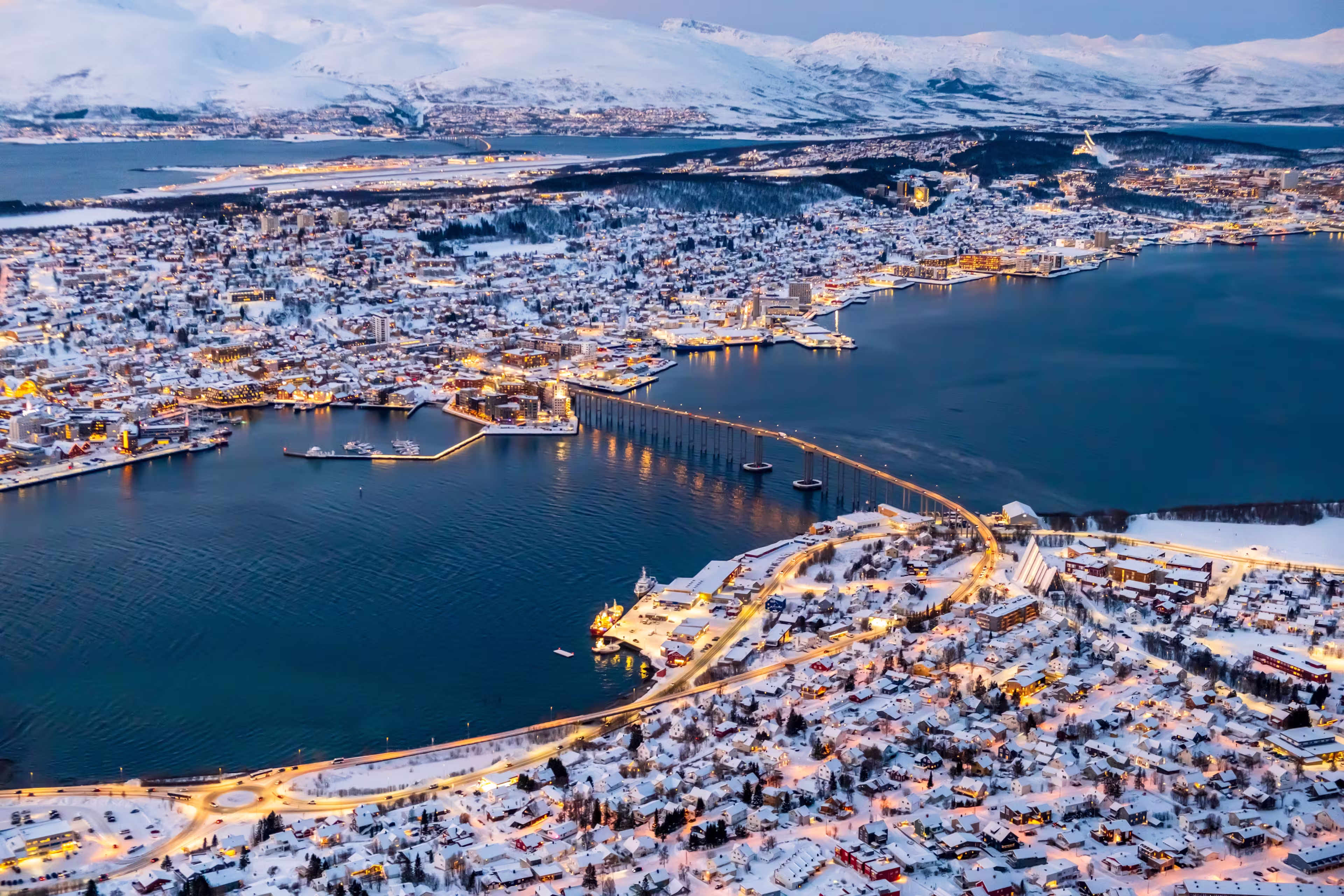 Aerial view of Tromso, Norway with city lights in winter from Storsteinen mountain. Tromsoya island with snowy roofs of Tromso town, harbor, Sandnessundet bridge over Fjord and mountains in background