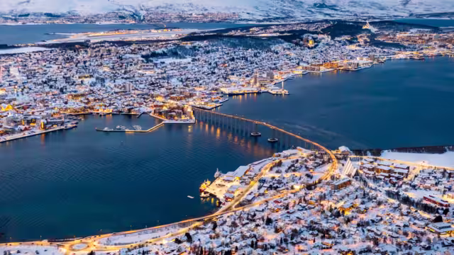 Aerial view of Tromso, Norway with city lights in winter from Storsteinen mountain. Tromsoya island with snowy roofs of Tromso town, harbor, Sandnessundet bridge over Fjord and mountains in background
