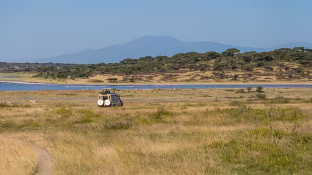 A safari vehicle makes its way across the golden plains of Ndutu, with Lake Ndutu and the distant highlands forming a dramatic backdrop to the wilderness.