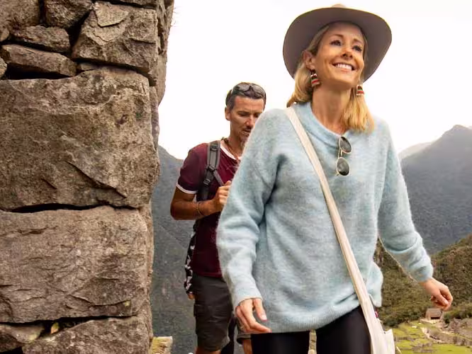 A blonde woman with a blue sweater walk in front of a man in a red t-shirt, with Machu Picchu in the background