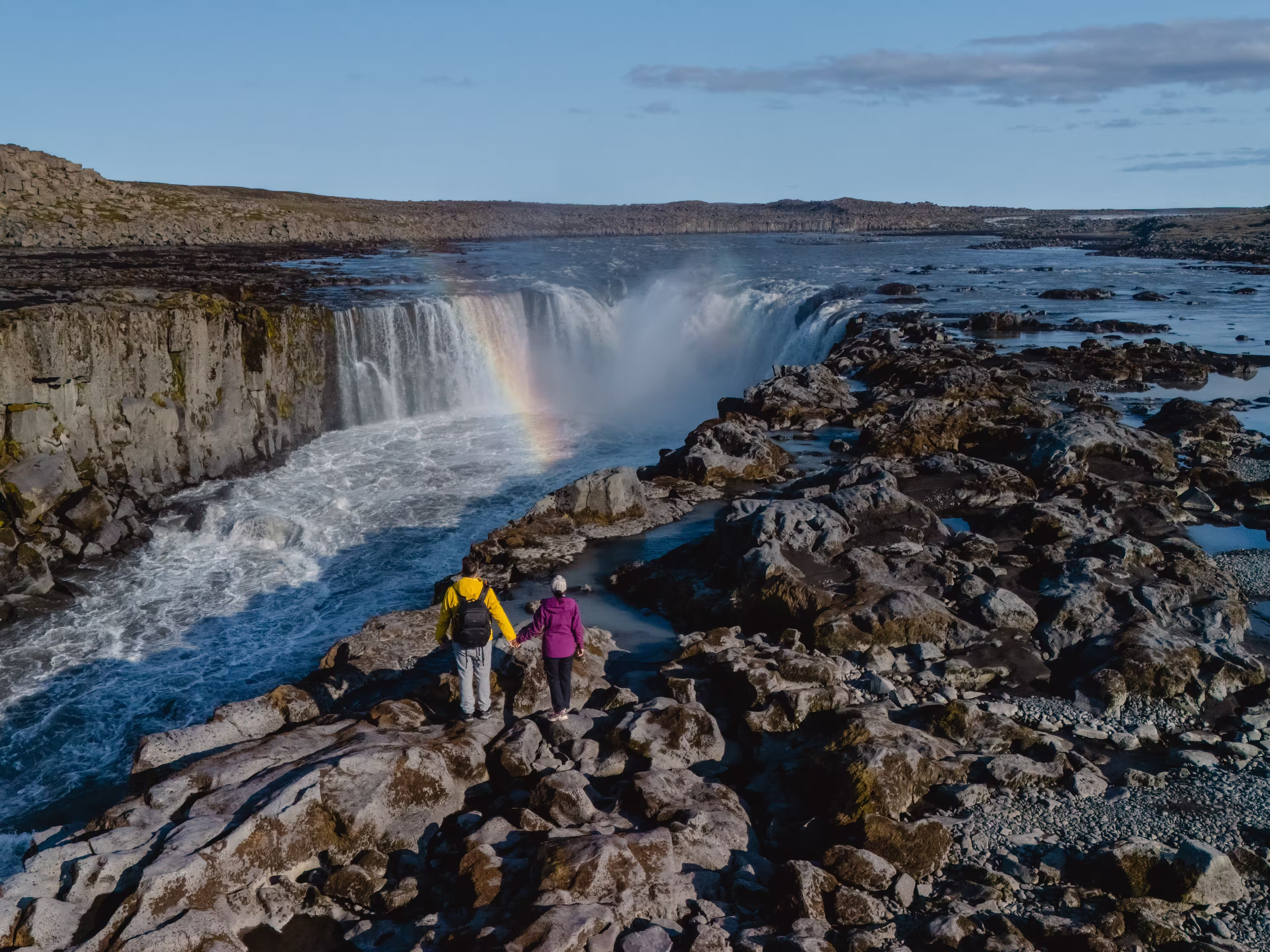 Visitors stand on rocky terrain, admiring the majestic Selfoss waterfall glistening under the sun in Iceland, A couple on vacation in Iceland