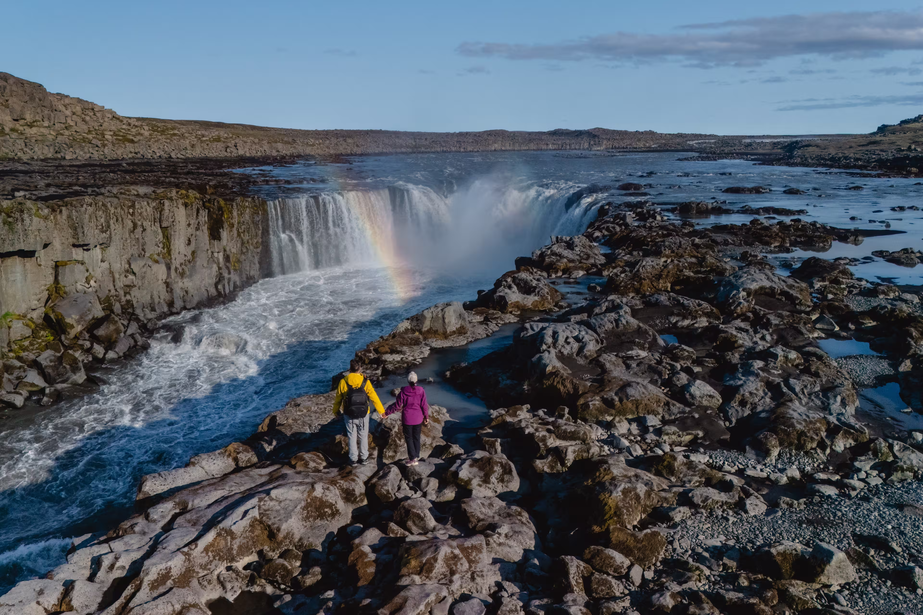 Visitors stand on rocky terrain, admiring the majestic Selfoss waterfall glistening under the sun in Iceland, A couple on vacation in Iceland