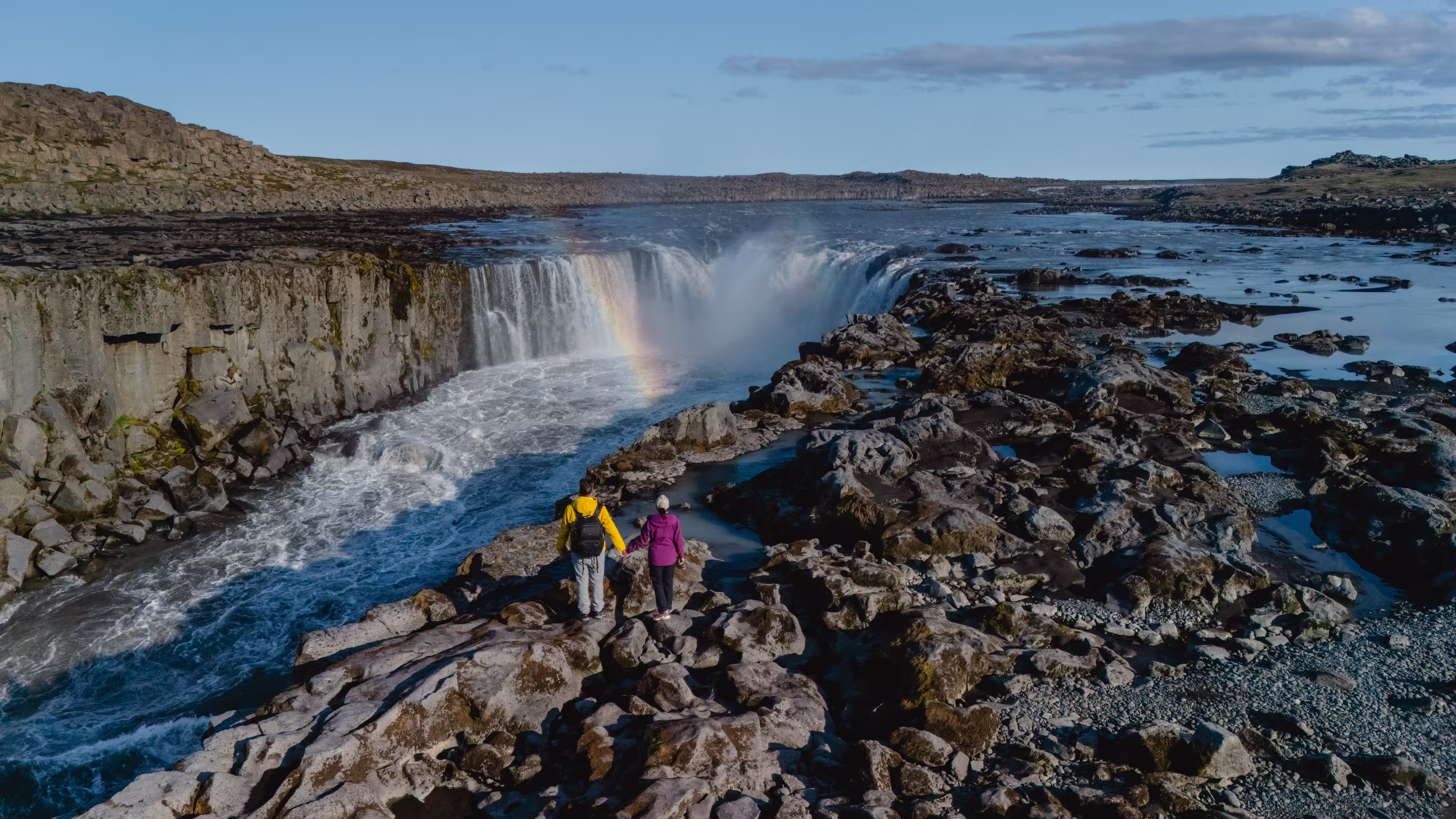 Visitors stand on rocky terrain, admiring the majestic Selfoss waterfall glistening under the sun in Iceland, A couple on vacation in Iceland