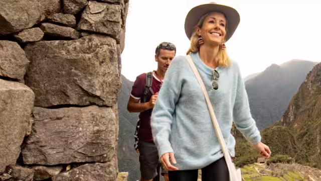 A blonde woman with a blue sweater walk in front of a man in a red t-shirt, with Machu Picchu in the background