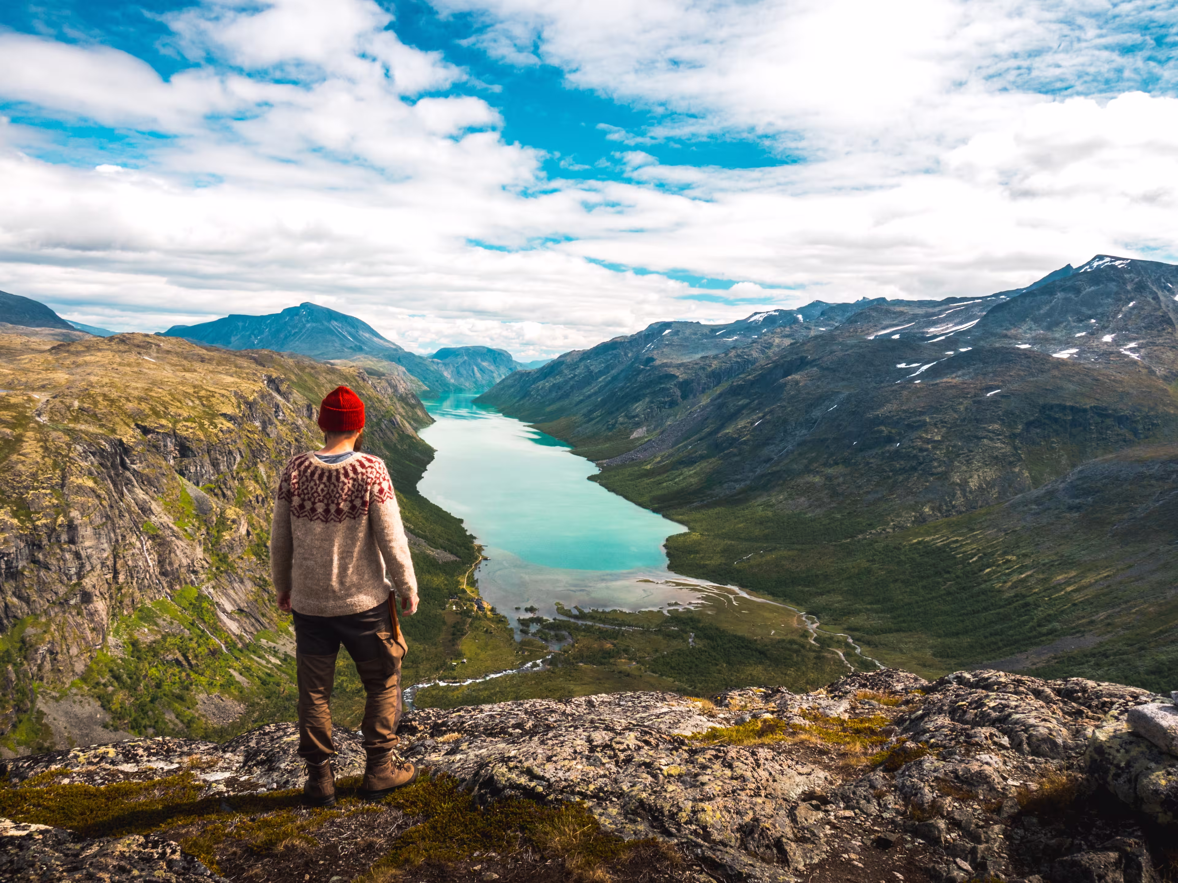 Hiker in red beanie and nordic sweater admires a scenic view from Gjendetunga peak over the lake of Gjende during beautiful autumn day in Jotunheimen national park, Norway