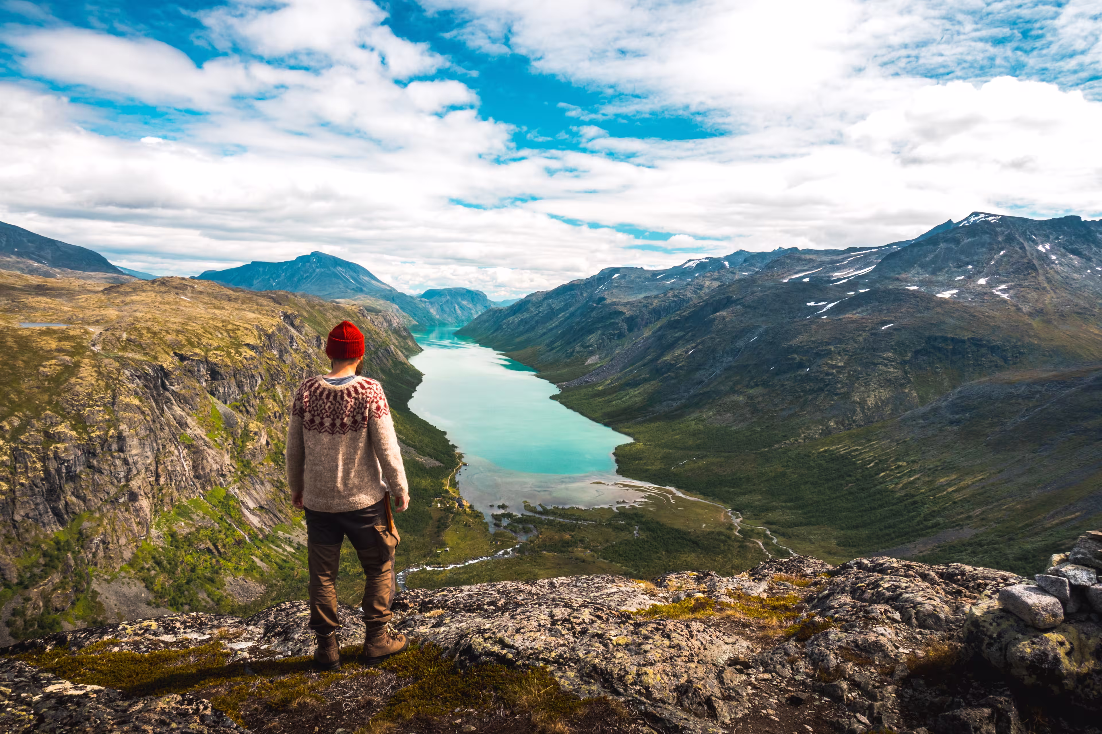 Hiker in red beanie and nordic sweater admires a scenic view from Gjendetunga peak over the lake of Gjende during beautiful autumn day in Jotunheimen national park, Norway