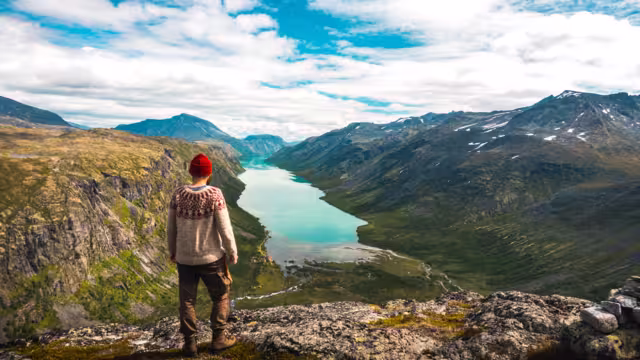 Hiker in red beanie and nordic sweater admires a scenic view from Gjendetunga peak over the lake of Gjende during beautiful autumn day in Jotunheimen national park, Norway