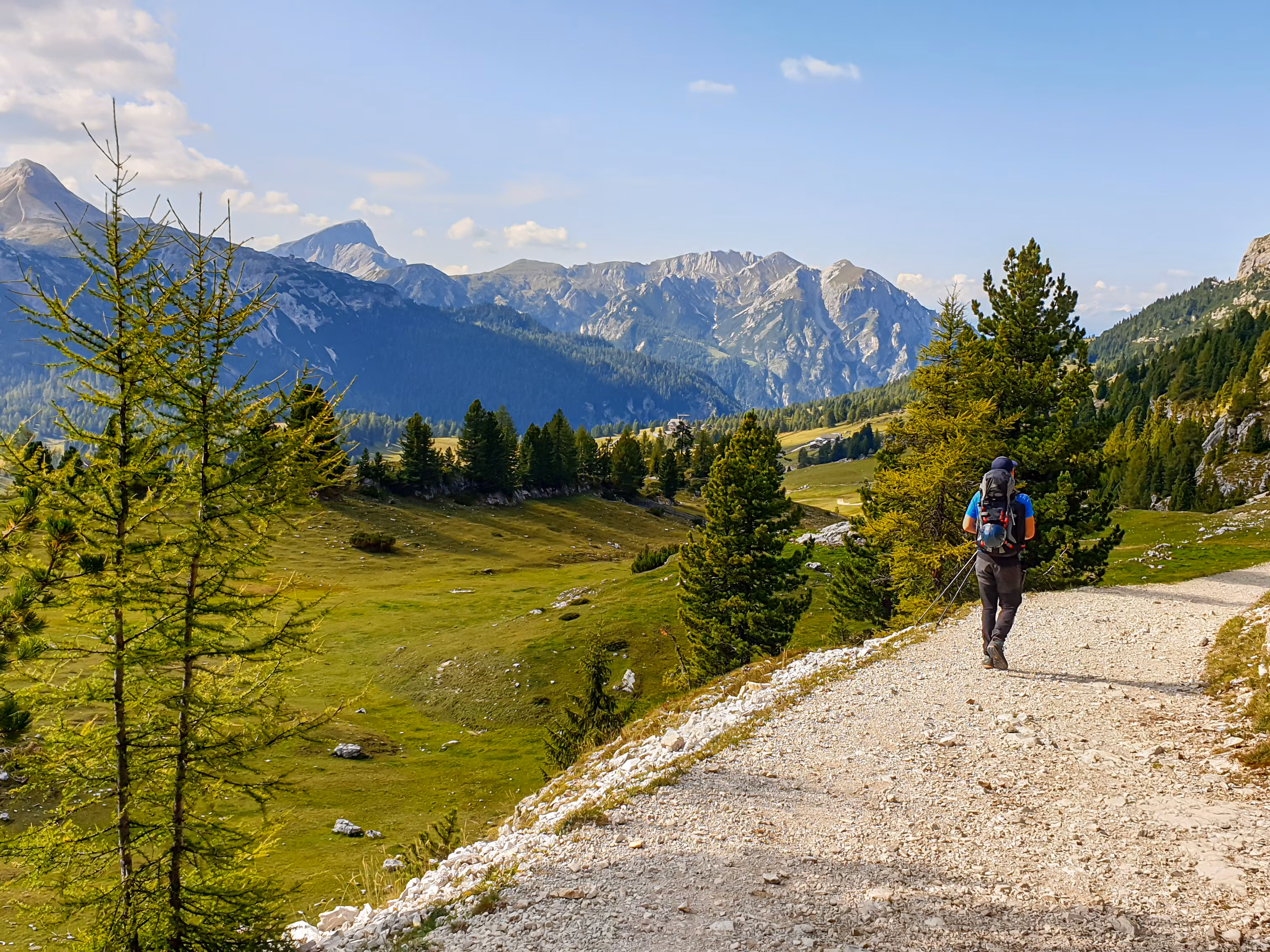 A man hiking along a wide gravelled road in Italian Dolomites from the top of Strudelkopf. A panoramic view on the high mountains. Sunny day. A few clouds above the high peaks. Lush green plateau