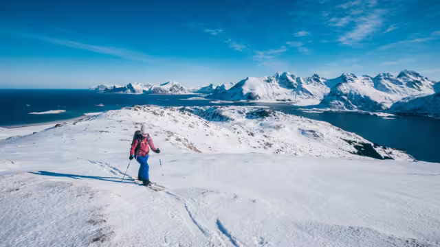 Girl skialper reaching the top after a long climb towering over the ocean and the peaks of Lofoten Island