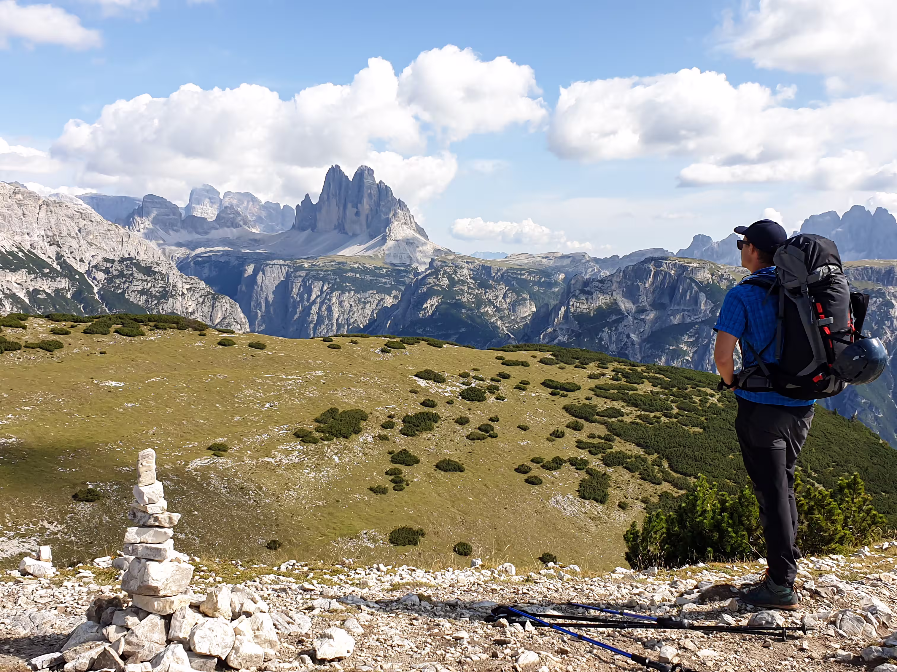 Man with a big hiking backpack and distant view on Drei Zinnen from Strudelkopf in Italian Dolomites. A lush green meadow in front. The man enjoys the view and the trek. High Alpine landscape. Freedom