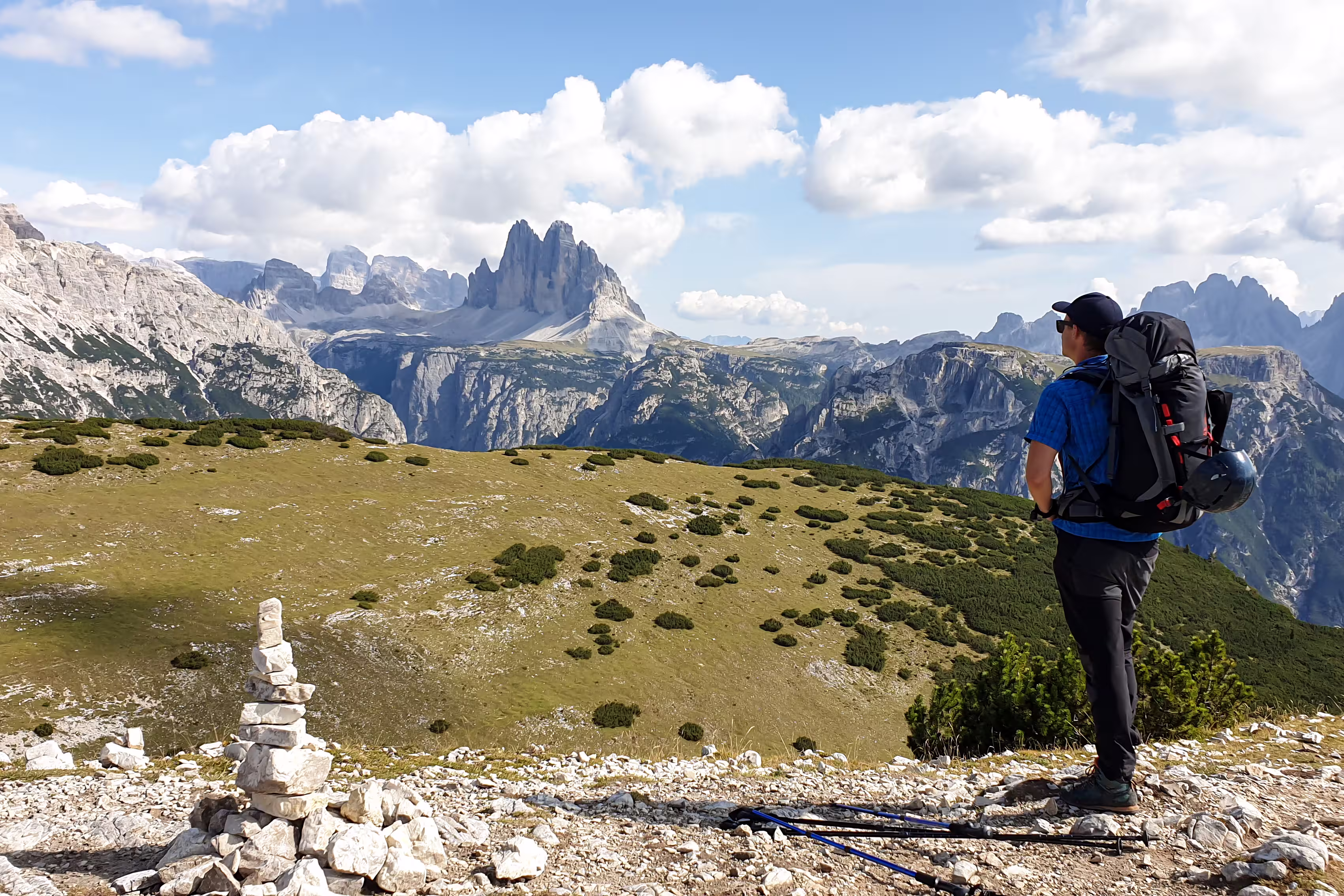 Man with a big hiking backpack and distant view on Drei Zinnen from Strudelkopf in Italian Dolomites. A lush green meadow in front. The man enjoys the view and the trek. High Alpine landscape. Freedom