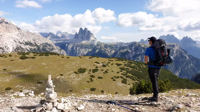 Man with a big hiking backpack and distant view on Drei Zinnen from Strudelkopf in Italian Dolomites. A lush green meadow in front. The man enjoys the view and the trek. High Alpine landscape. Freedom