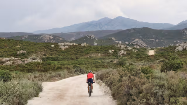 Cycling. Offroad Cycling in the Agriates Desert. Corsica, France. a red shirt cyclist, Dusty road, Rocky road, Mediterranean Vegetation. Mountains, Daylight. Cloudy sky. Courage for the Journey.